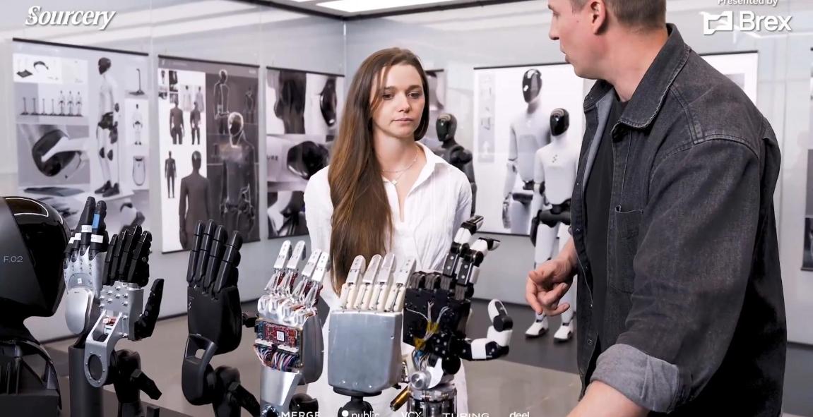 Successive generations of Figure robot hands displayed on stands as Brett Adcock explains them