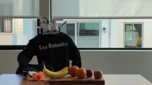 Humanoid robot in a black Tau Robotics branded shirt at a dining table, with a white sensor head and robotic gripper hand clearly visible