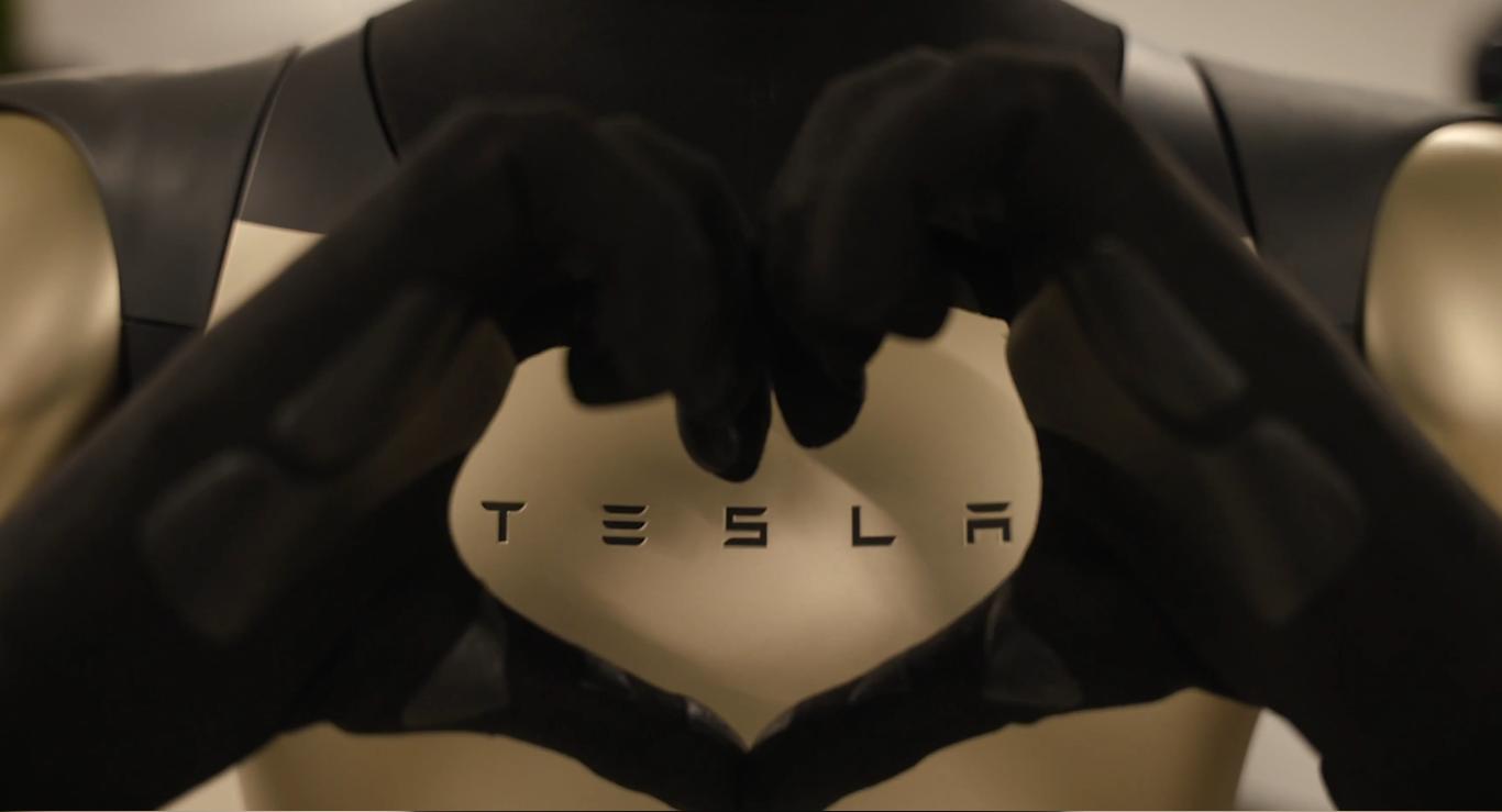 Close-up of two Tesla Optimus robot hands forming a heart shape over the Tesla logo on the robot chest